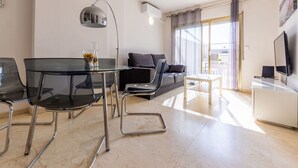 Een moderne woonkamer met een glazen eettafel, zwarte stoelen, een grijze bank, een witte salontafel en een televisie op een witte kast.