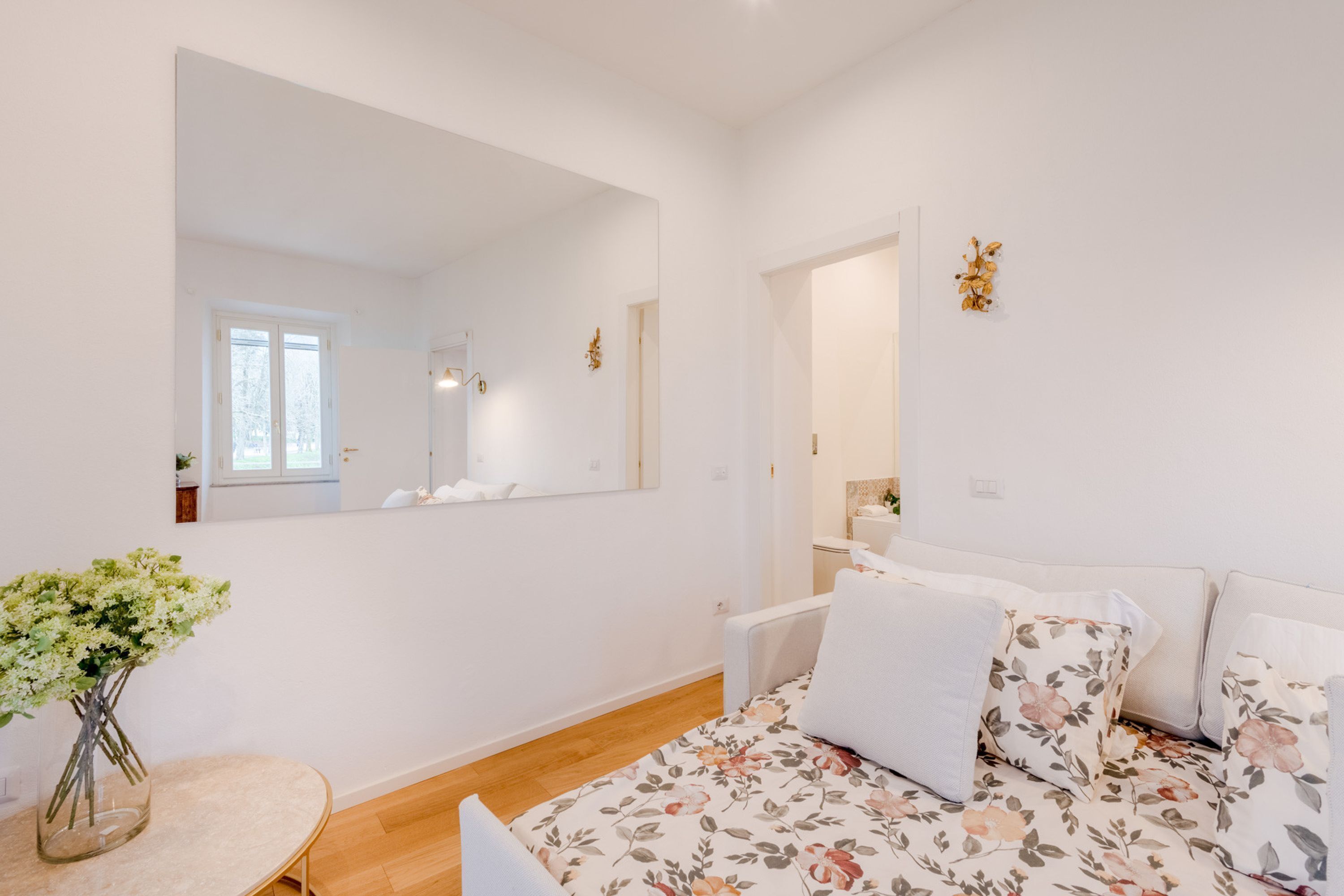 Une chambre à coucher moderne avec un grand miroir, une couvre-lit à motifs floraux et un vase de fleurs posé sur une table ronde.