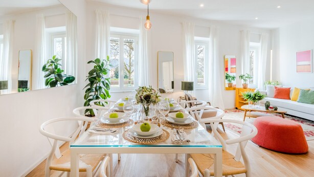 Un comedor moderno con una mesa de vidrio, sillas blancas y un otomano rojo.