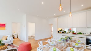 Una cocina moderna con armarios blancos, una zona de comedor con una mesa de vidrio dispuesta para seis personas y un otomano rojo.