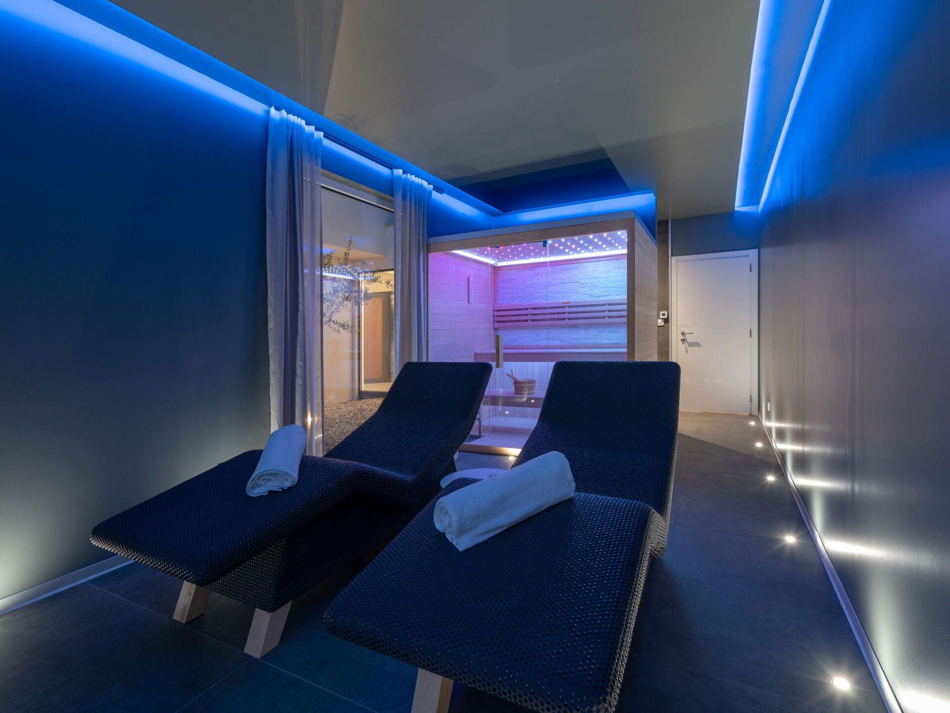 Une salle de spa dotée de deux fauteuils de couleur sombre, d’une porte en verre donnant accès à une zone de soins et d’un éclairage d’ambiance.