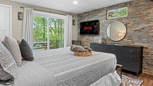 A bedroom with a large bed, a TV mounted on a stone wall, a dresser, and a balcony with a view of trees. - Walk to Town-Lake Views-6 Decks-Pool Table-FirePit: The Lookout (Lake Harmony)