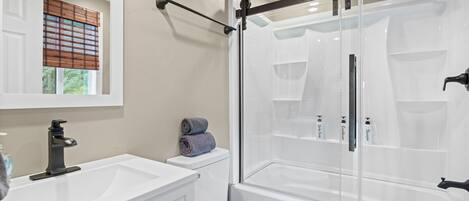 A modern bathroom with a glass-enclosed shower, white fixtures, and a window with blinds.