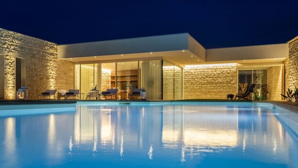 A modern house with a swimming pool, stone walls, and a glass-enclosed patio area with outdoor furniture.