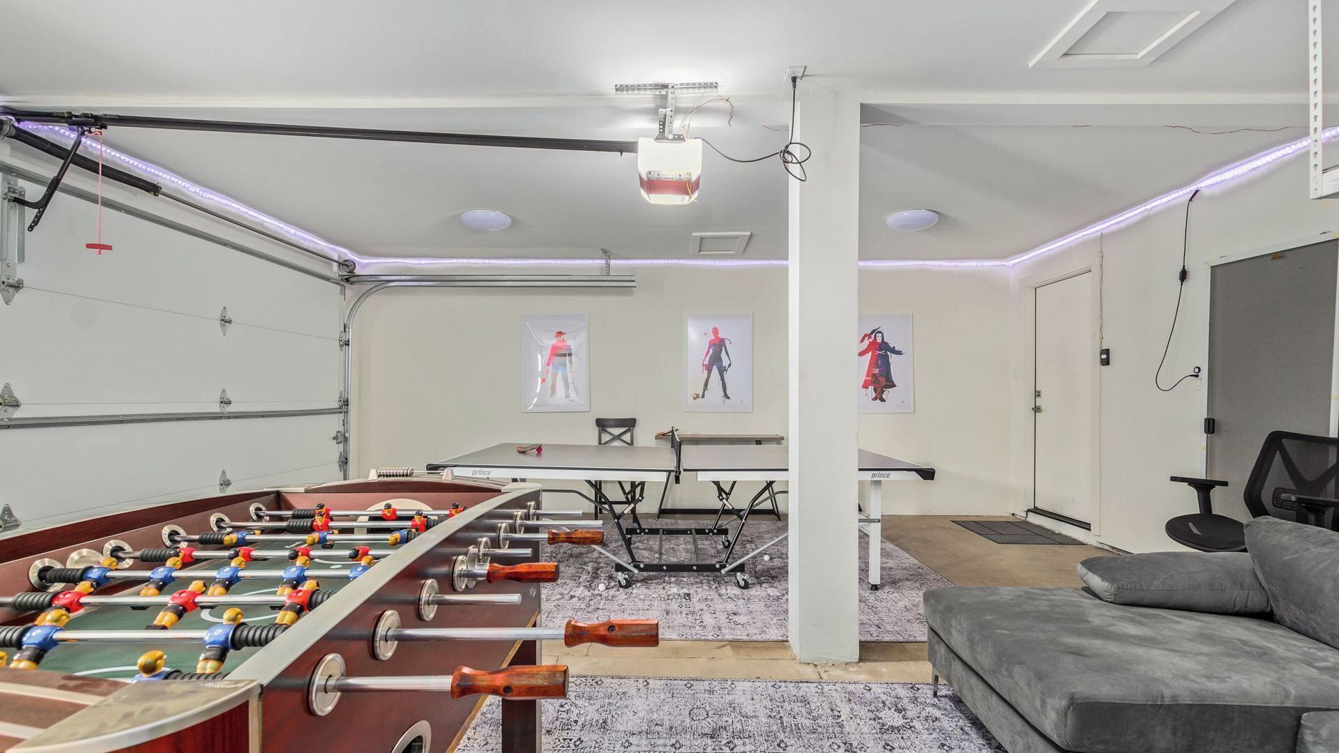 Ein Zimmer mit Tischfußball, einem grauen Sofa, einem Schreibtisch mit Computer und Kunstwerken an den Wänden.