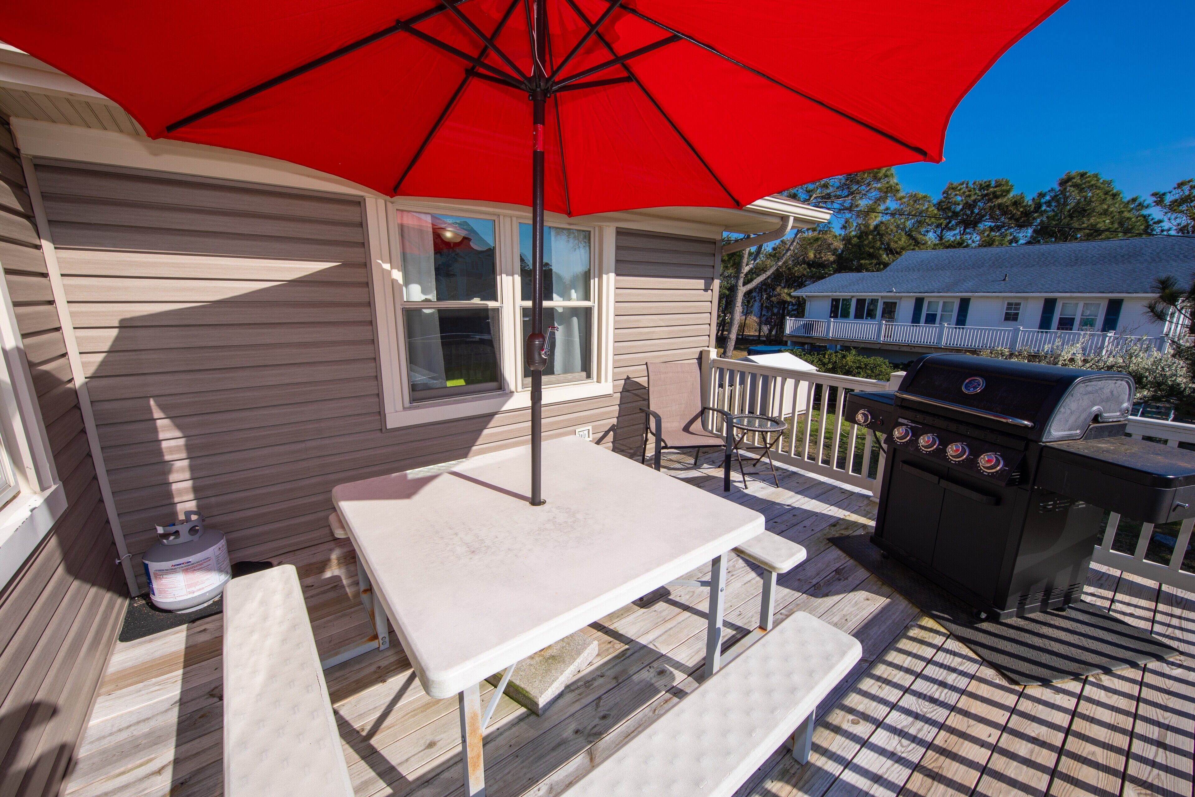 A wooden deck with a white table and benches, a red umbrella, and a barbecue grill.