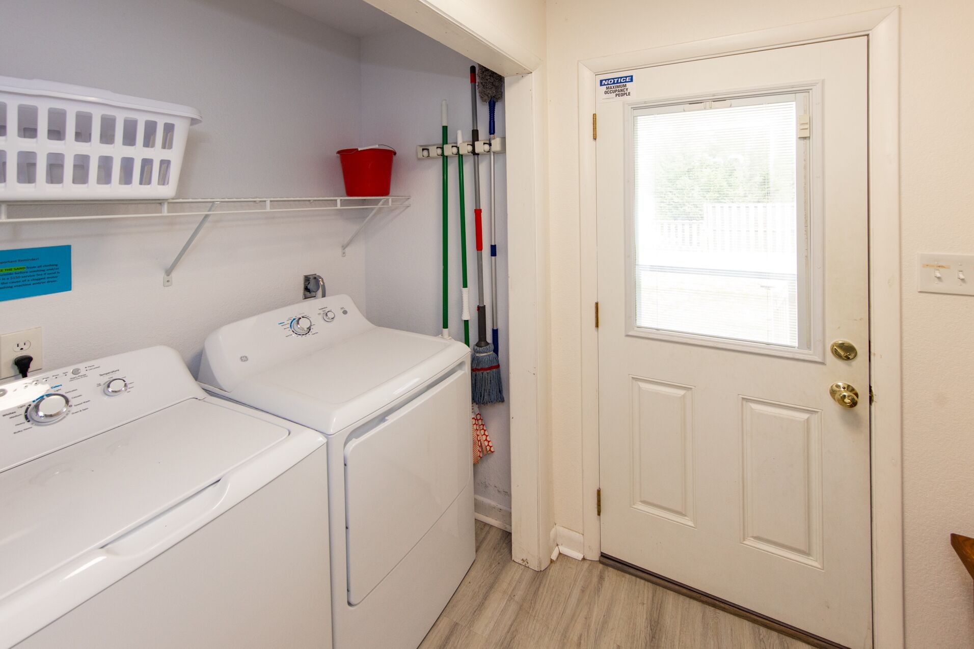 A laundry room with a washing machine and dryer, a shelf with a basket, and a door with a maximum occupancy sign.
