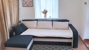 Un salon chaleureux avec un canapé d’angle, une fenêtre ornée de rideaux légers et une œuvre d’art murale aux motifs floraux.