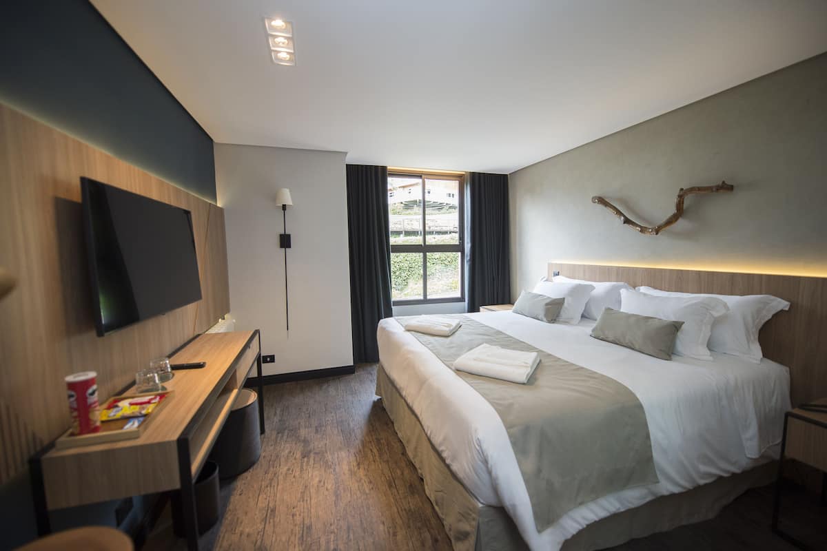 A modern hotel room with a large bed, a flat-screen TV mounted on the wall, a desk with a chair, and a window providing a view of greenery outside.