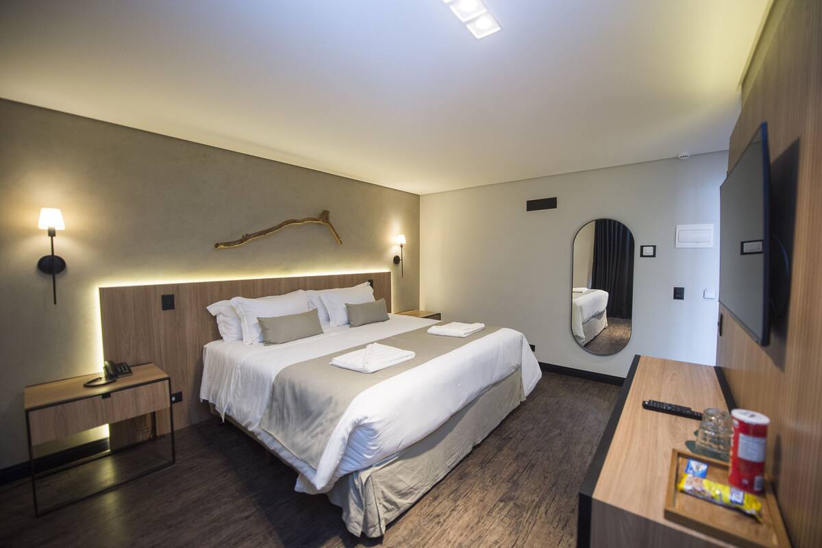 A hotel room with a large bed, bedside tables, a desk with a chair, a mirror, and a flat-screen TV.