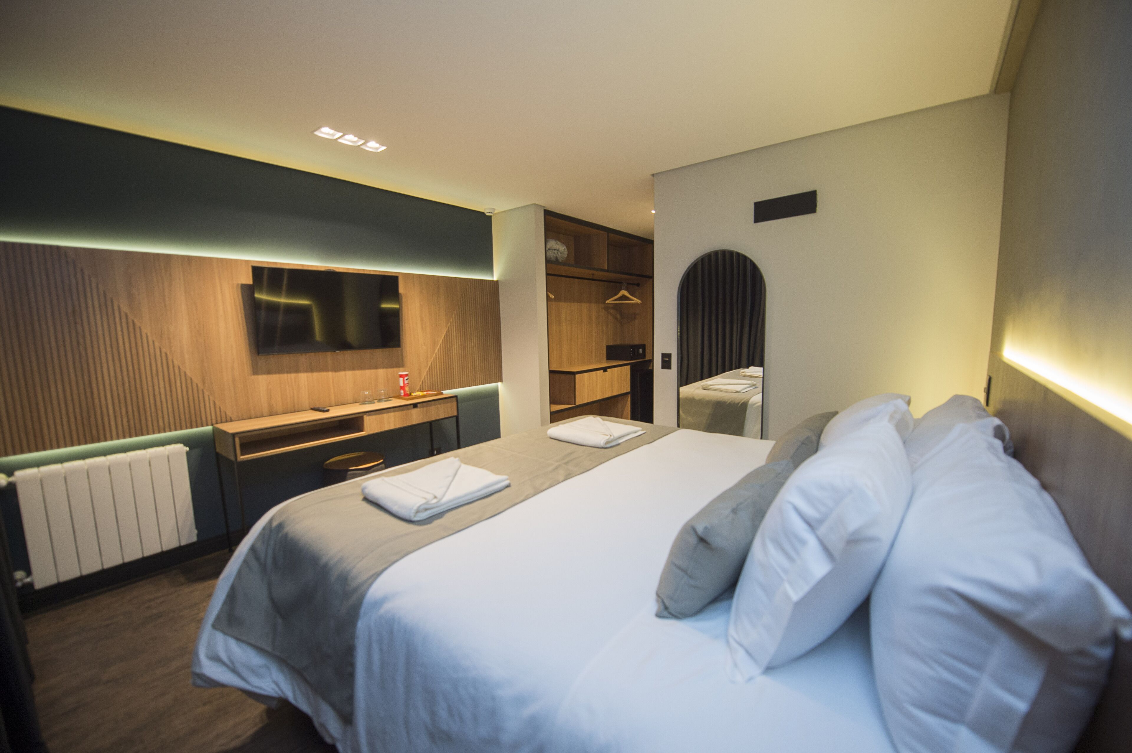 Quarto de hotel moderno com uma cama bem arrumada, uma escrivaninha de madeira, uma televisão de tela plana, um closet espaçoso e um radiador.
