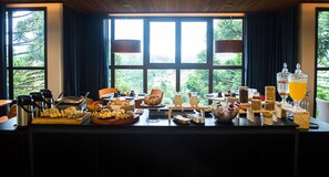 A breakfast buffet with various food items and beverages, set against a backdrop of large windows with a view of greenery. - Pinot Hotel (Campos do Jordão)