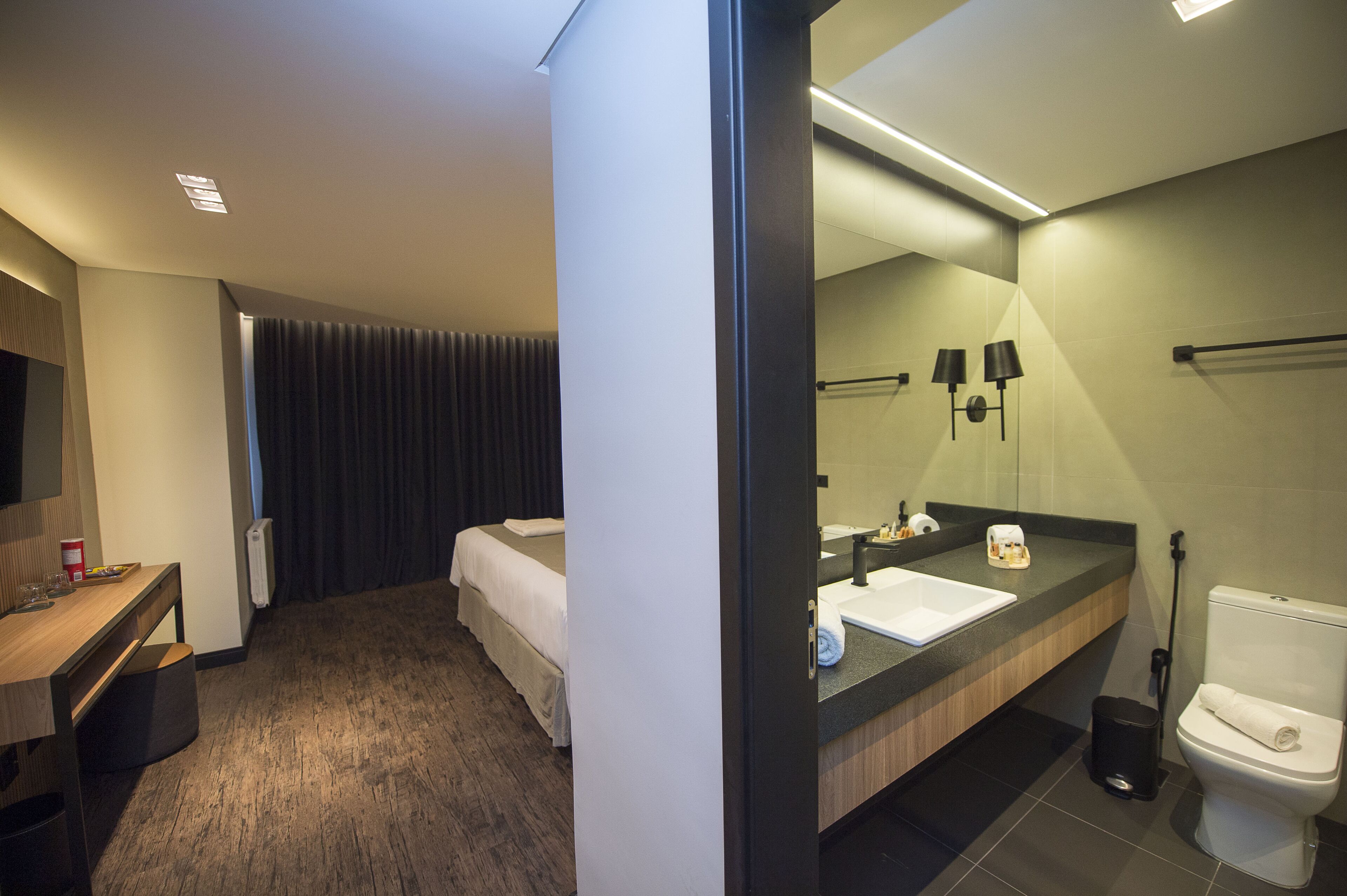 Habitación de hotel con cama, escritorio con silla, baño con lavamanos, espejo y retrete.