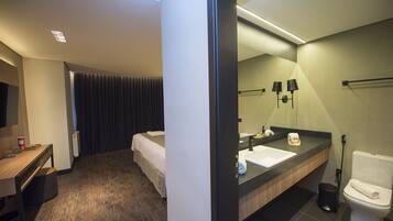 Habitación de hotel con cama, escritorio con silla, baño con lavamanos, espejo y retrete.