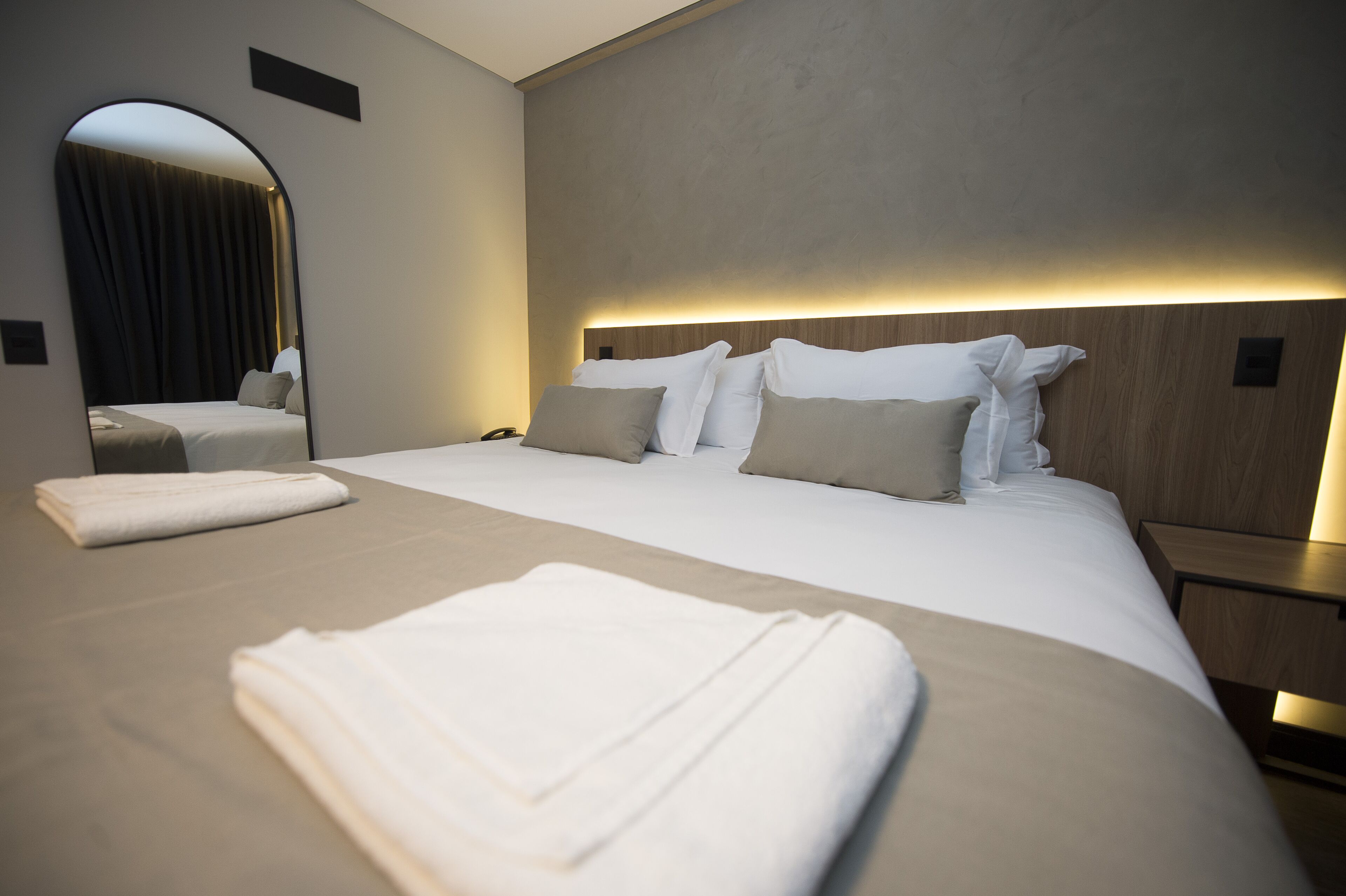 Une chambre d’hôtel moderne dotée d’un grand lit, d’une table de chevet, d’un miroir et d’une tête de lit avec un éclairage intégré.