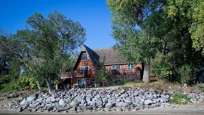 A rustic cabin with a stone foundation, large windows, and a balcony overlooking a rocky shoreline. - Lakefront Lake Poinsett Cottage with Private Beach (Castlewood)