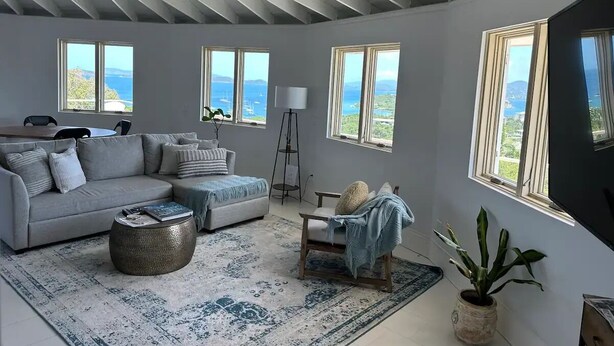 A spacious living room with a grey sofa, a coffee table, and a view of the sea through large windows. - The Round House (St Thomas)