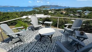 A balcony with Adirondack chairs and a small table, overlooking a marina with boats and a coastal landscape. - The Round House (St Thomas)