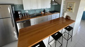 A modern kitchen with white cabinets, a wooden countertop, a stainless steel refrigerator, a gas stove, and a wooden ceiling beam. - The Round House (St Thomas)