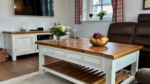 Une table à manger avec un saladier de fruits et des verres à vin, un téléviseur au-dessus, et un canapé en cuir marron en arrière-plan.