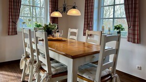 Un coin repas avec une table en bois et des chaises blanches, deux lampes suspendues et des rideaux à carreaux.