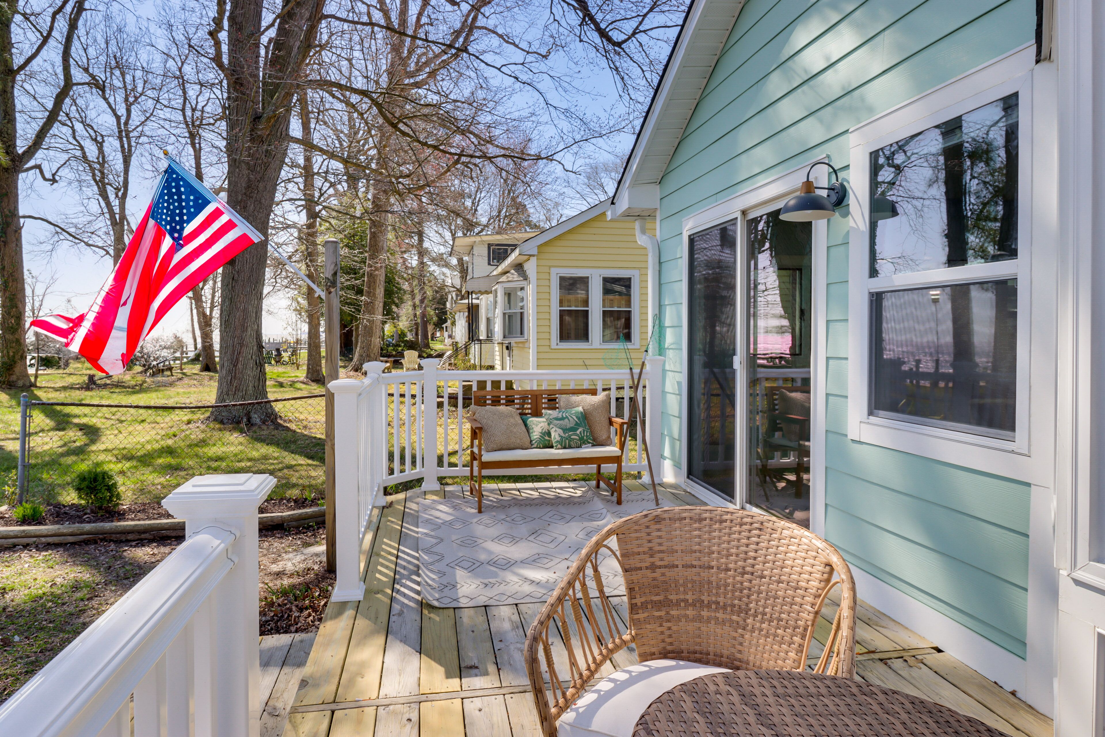 A wooden deck with a wicker chair and a bench, a white fence, and a house with large windows.