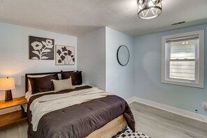 Ein Schlafzimmer mit Bett, Nachttisch, Lampe, Wandbildern, einem Spiegel und einem Fenster.