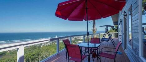 A wooden deck with a table and chairs, a red umbrella, and a view of the ocean.