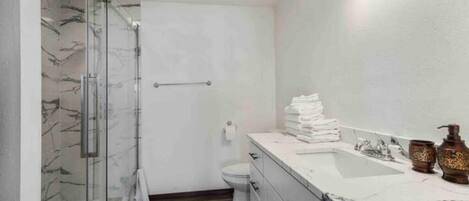 Un baño moderno con cabina de ducha de vidrio, inodoro blanco, lavabo con azulejos de mármol y tocador con espejo.