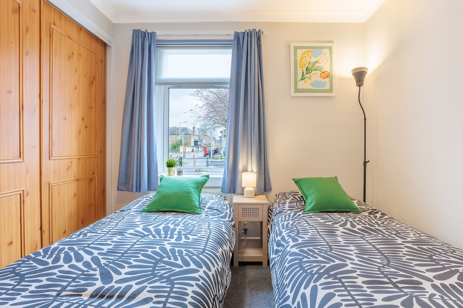 Een kamer met twee bedden, een raam met gordijnen, een lamp en een schilderij aan de muur.