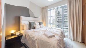 Une chambre d’hôtel moderne avec un grand lit, une table de chevet avec une lampe, une vue sur la ville grâce à de grandes fenêtres et un panorama urbain visible à l’extérieur.