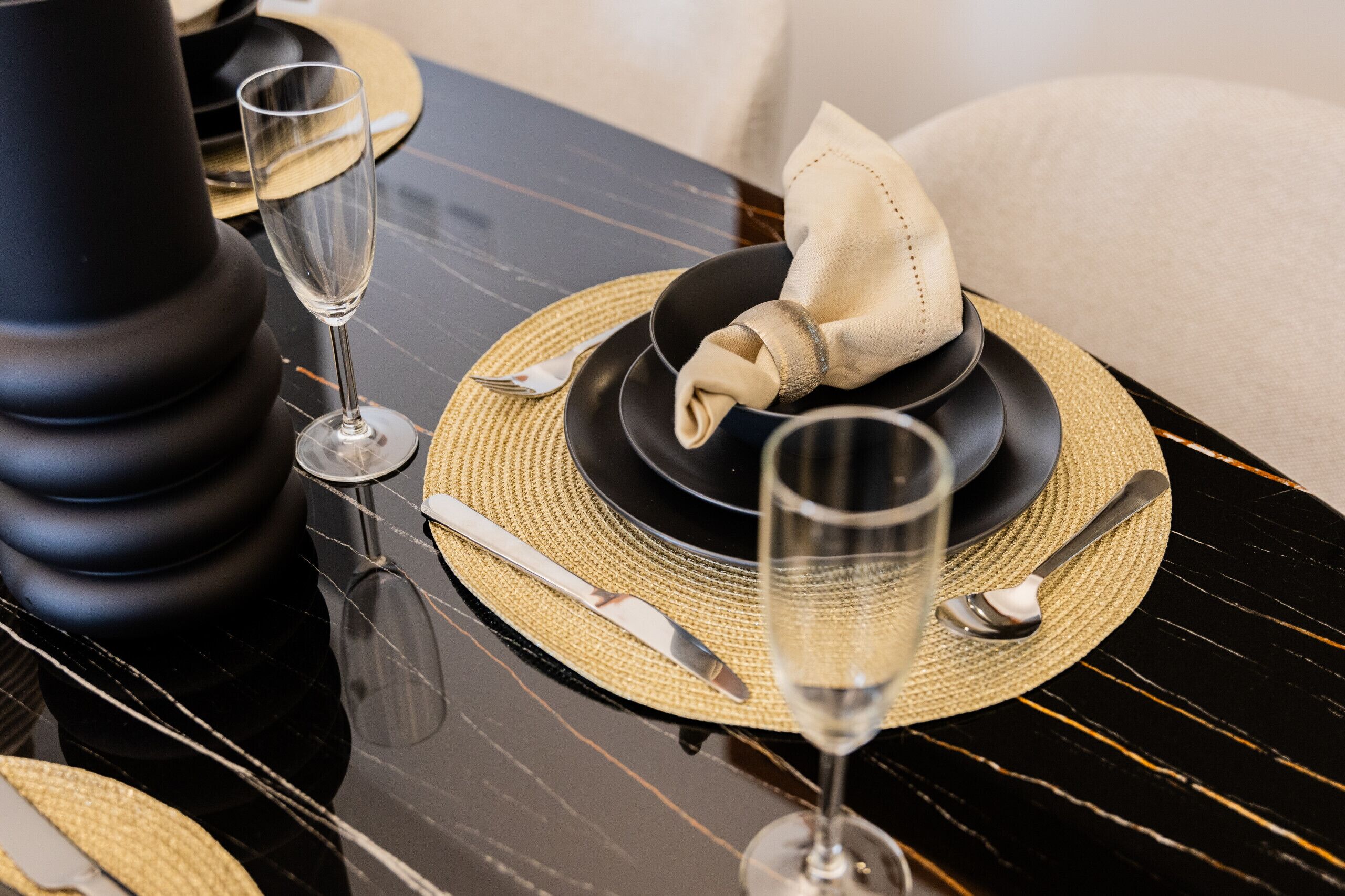 Uma mesa de jantar posta com pratos pretos, guardanapos dourados e bege, e copos de vinho transparentes.