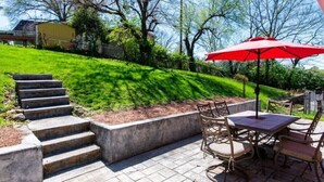 Eine Terrasse mit Tisch und Stühlen, einem roten Sonnenschirm und einer Treppe aus Beton, die einen grasbewachsenen Hang hinaufführt.
