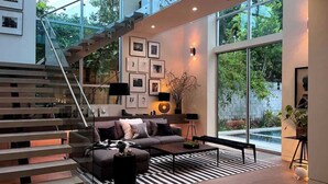 A modern living room with a staircase, a sofa, a coffee table, and a large window offering a view of greenery. - Stunning Home: Pool, Hot Tub, Fire Pit, Dogs OK (Los Angeles)