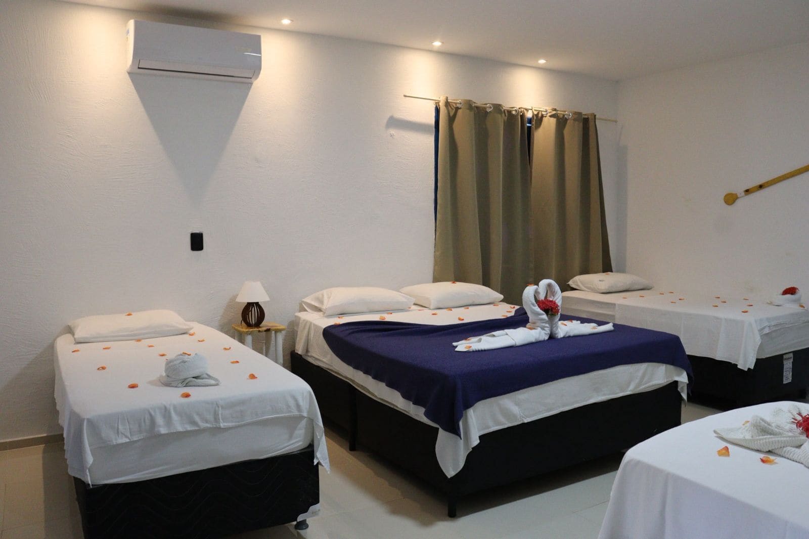 A hotel room with three beds, each with a white bedspread and pillows, a bedside table with a lamp, and a wall-mounted air conditioner.