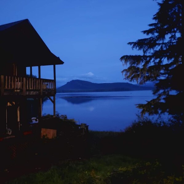 A wooden cabin with a balcony overlooking a calm lake and distant mountains.