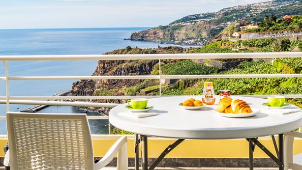 A table with breakfast items and a chair on a balcony overlooking a coastal view. - Cantinho do Sol by Homie (Ribeira Brava)