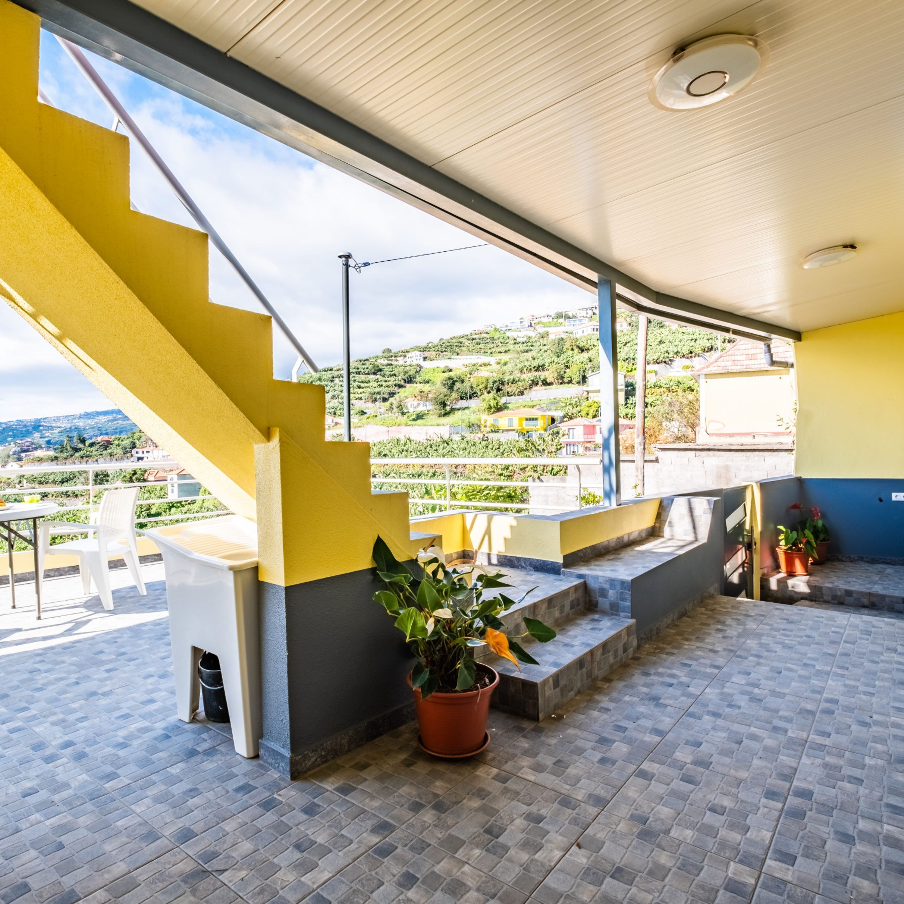 Un espace extérieur abrité, doté d’un escalier jaune, de plantes en pot et offrant une vue sur une colline.