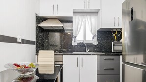 A modern kitchen with white cabinets, a stainless steel refrigerator, a black countertop, and a window with curtains. - Cantinho do Sol by Homie (Ribeira Brava)