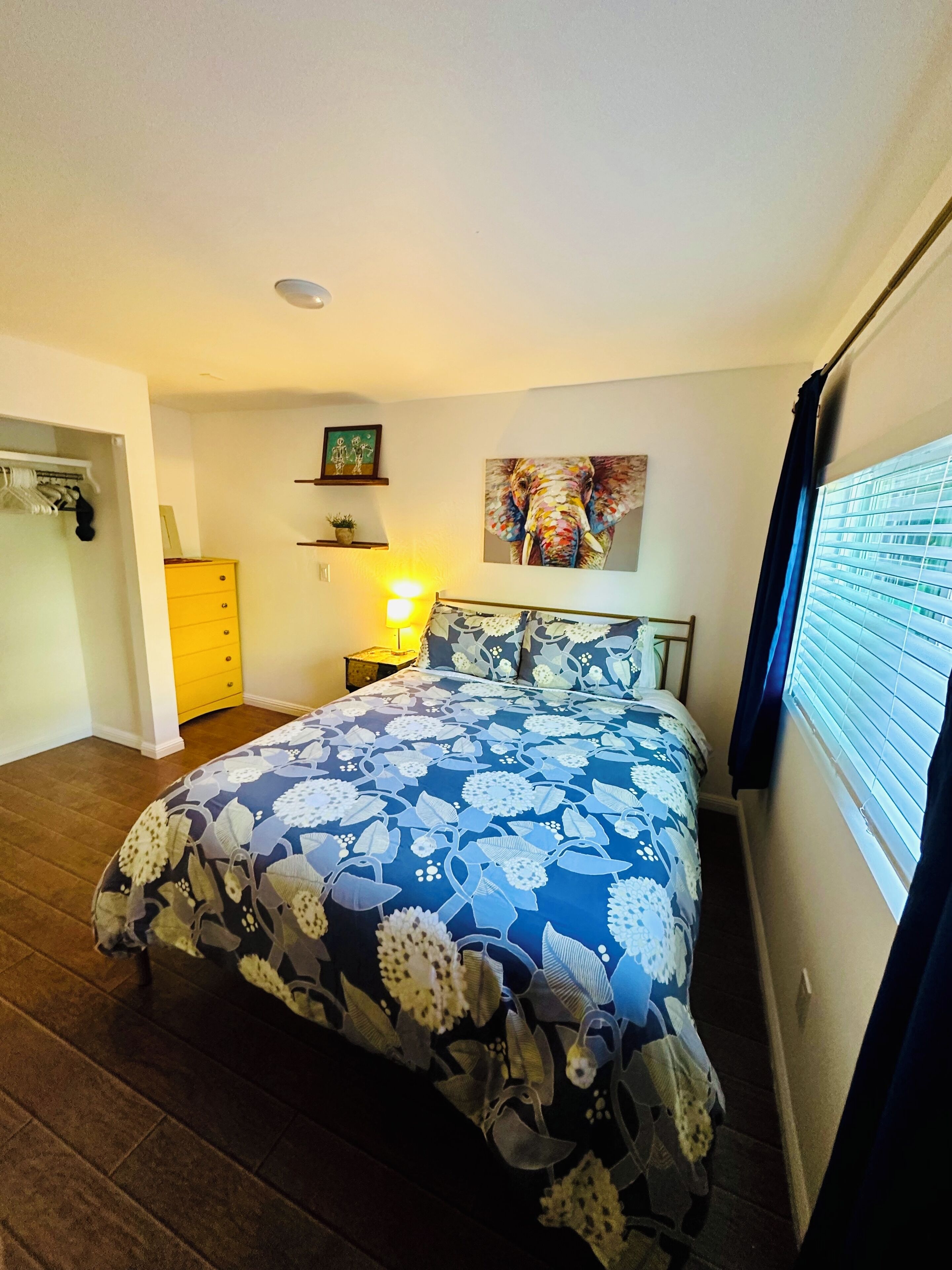 A bedroom with a floral bedspread, a nightstand with a lamp, a painting of an elephant, and a window with blinds.