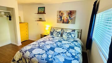 A bedroom with a floral bedspread, a nightstand with a lamp, a painting of an elephant, and a window with blinds.