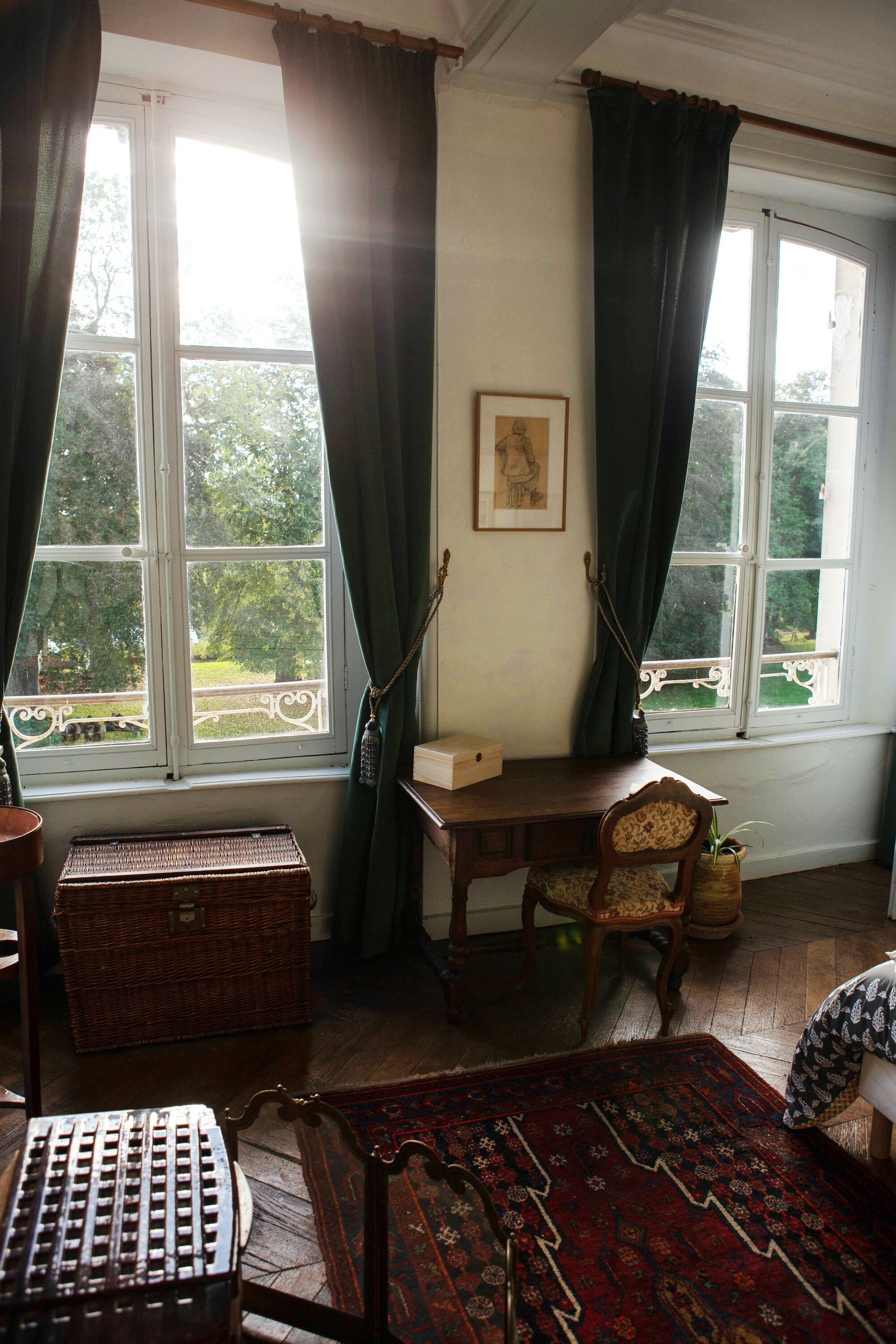 A room with a large window, a wooden desk, a chair, a wicker chest, and a patterned rug.