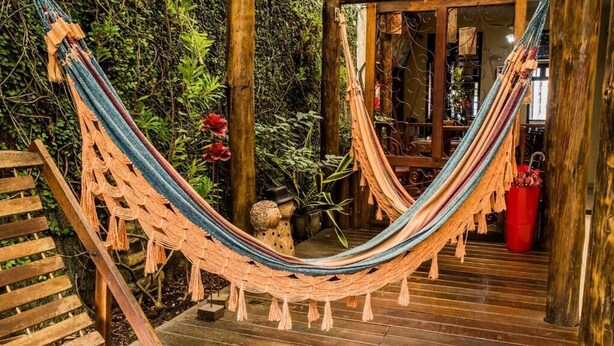 A hammock suspended between wooden posts in a garden area with a wooden bench and a red bucket. - Baobá Pousada e SPA (Olinda)