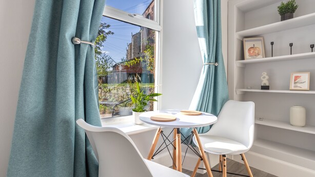 A modern room with a white chair, a small round table, and a window with teal curtains. - Beautiful Ground Floor Apartment (Perth)