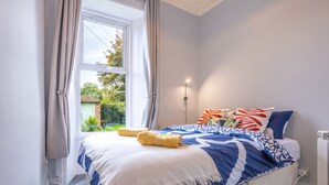 A neatly made bed with a blue and white patterned bedspread, a white throw blanket, and two yellow towels. There is a window with curtains, a wall-mounted light, and a radiator. - Beautiful Ground Floor Apartment (Perth)