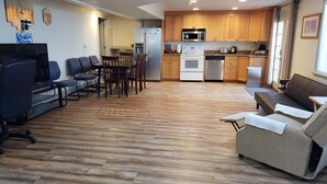A modern living room with a sofa, TV, and chairs. A kitchen with wooden cabinets and appliances is visible in the background. - 3 Bedroom Basement Suite with Kitchen (Centerville)