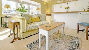 A cozy living room with a sofa, a coffee table, a side table, and a chair. There is a window with curtains, a lamp, and a shelf with decorative items.