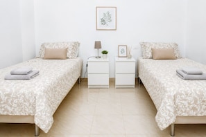 A twin room with two beds, each with a matching pillow and a folded towel. There is a nightstand with a lamp, a framed picture, and a small plant. The room has a white wall and a framed botanical print above the nightstand.