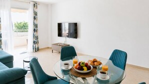 A modern living room with a glass dining table, teal chairs, and a flat-screen TV mounted on the wall.