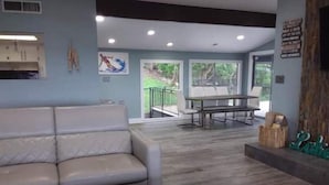 A modern living room with a white leather sofa, a black ottoman, and a dining table with chairs. There are large windows and a view of greenery outside. - Pet Friendly- Luxury home- Amazing Views-Lake Travis-25min Downtown Austin (Lakeway)